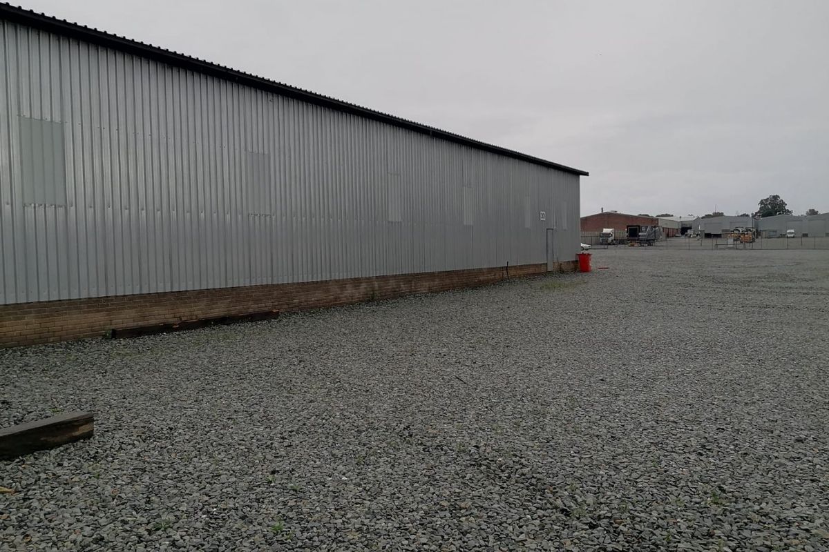 Rear view of an industrial warehouse building featuring corrugated metal cladding, a brick base, and a large gravel yard area.