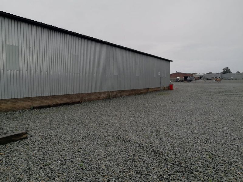 Rear view of an industrial warehouse building featuring corrugated metal cladding, a brick base, and a large gravel yard area.