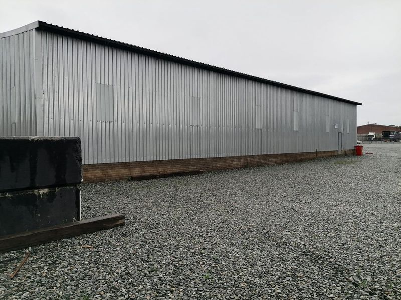 Exterior view of an industrial warehouse building featuring corrugated metal cladding, a brick base, and a spacious gravel yard area.