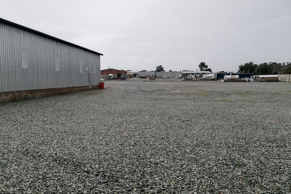 A large industrial yard featuring a corrugated metal warehouse building and extensive gravel surfacing, suitable for storage or logistics operations.