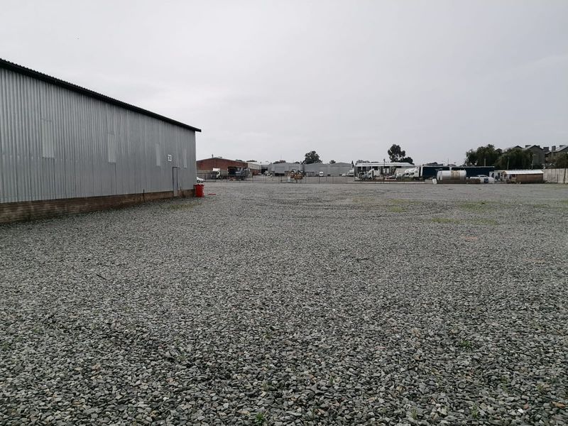 A large industrial yard featuring a corrugated metal warehouse building and extensive gravel surfacing, suitable for storage or logistics operations.