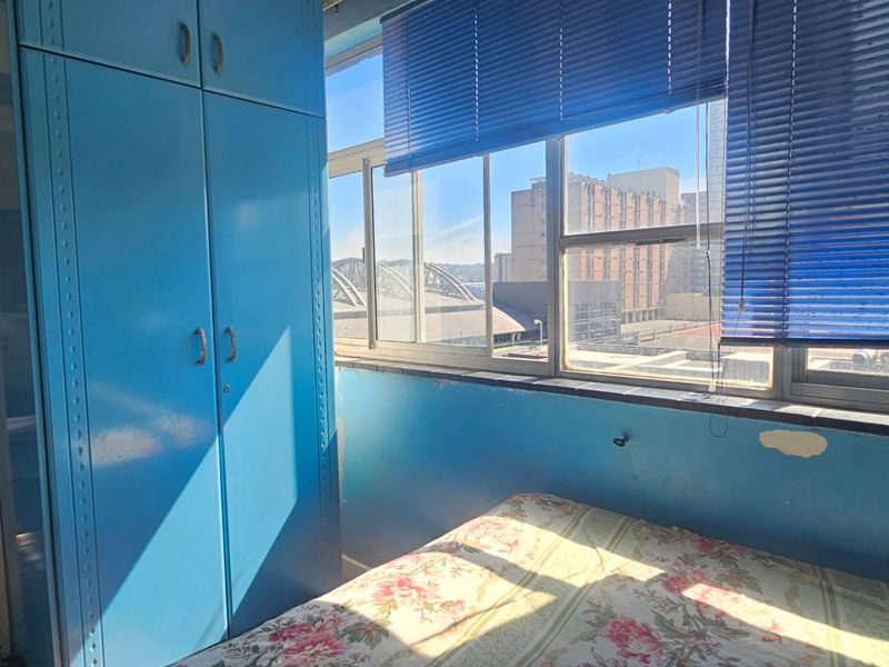 A bedroom featuring a large window with blue venetian blinds and a built-in blue wardrobe, offering an urban city view.