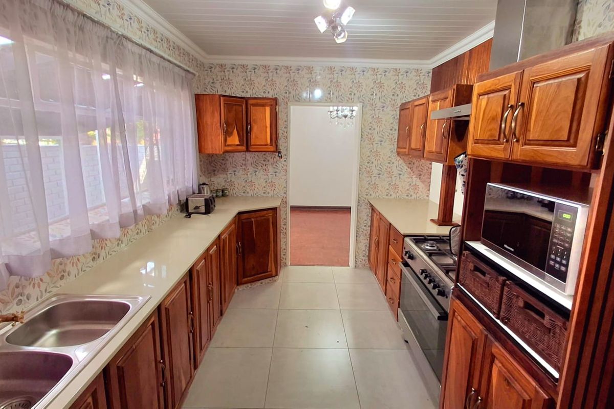 A functional kitchen featuring traditional wooden cabinetry, a gas hob with extractor, and double stainless steel sink, finished with tiled flooring and patterned wallpaper.