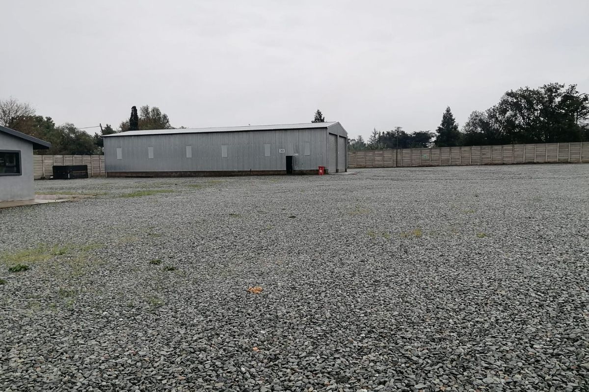 Industrial property featuring a large corrugated metal warehouse building set within a spacious, gravel-surfaced yard enclosed by a precast concrete boundary wall.