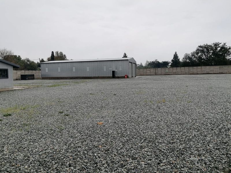 Industrial property featuring a large corrugated metal warehouse building set within a spacious, gravel-surfaced yard enclosed by a precast concrete boundary wall.