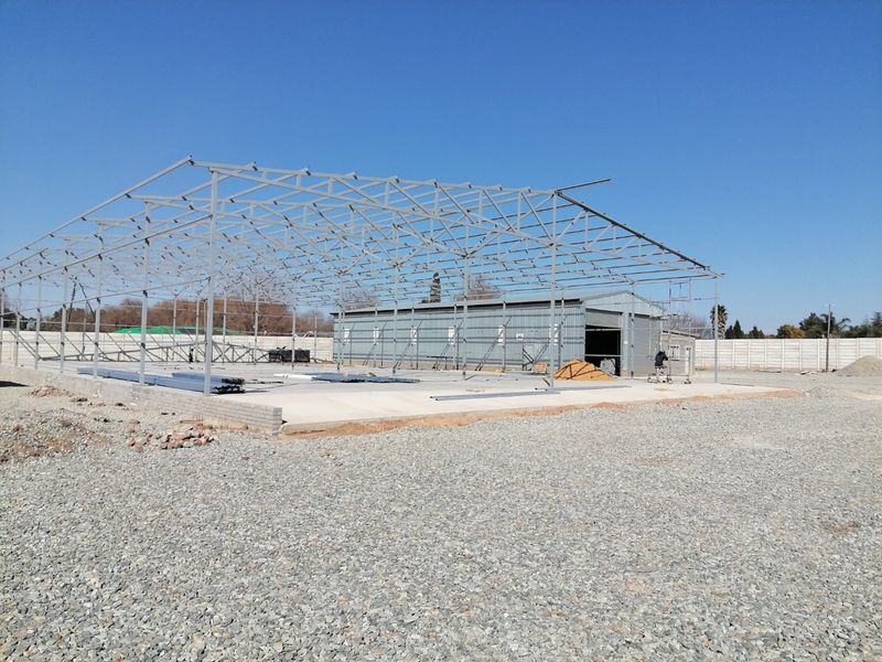 Industrial property under construction featuring a large steel frame structure, concrete slab foundation, and existing corrugated metal workshop building.