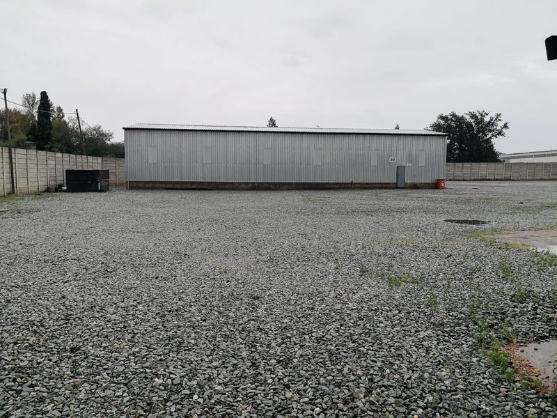 Industrial property featuring a large corrugated metal warehouse building situated on a spacious gravel yard enclosed by precast concrete boundary walls.