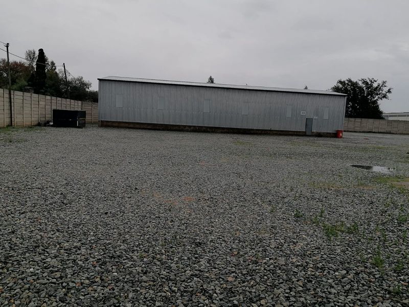 Industrial property featuring a large corrugated metal warehouse building set within a spacious gravel-surfaced yard enclosed by precast concrete boundary walls.