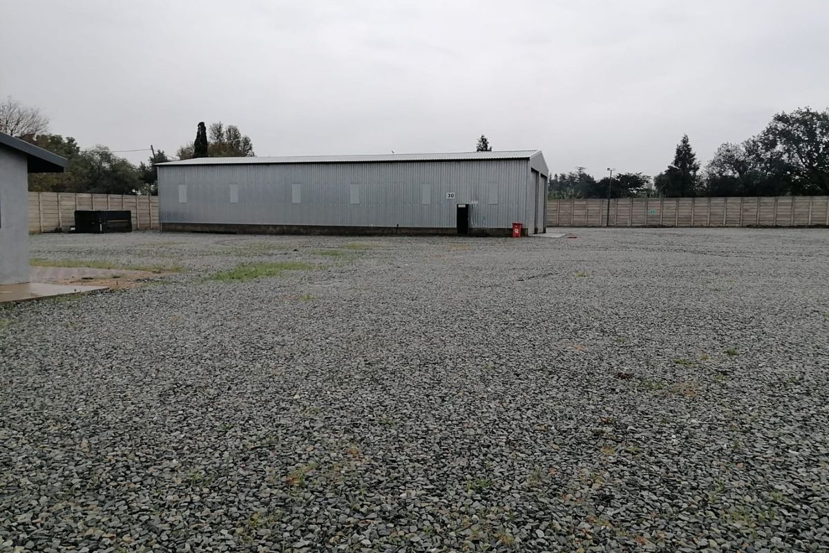 A spacious industrial yard featuring a corrugated metal warehouse building, enclosed by a precast concrete boundary wall with a gravel-surfaced yard area.