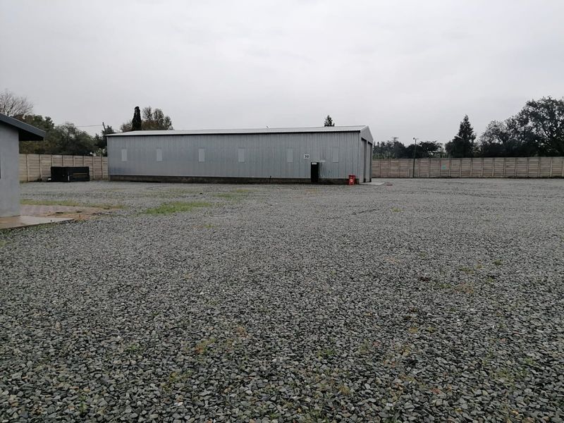 A spacious industrial yard featuring a corrugated metal warehouse building, enclosed by a precast concrete boundary wall with a gravel-surfaced yard area.