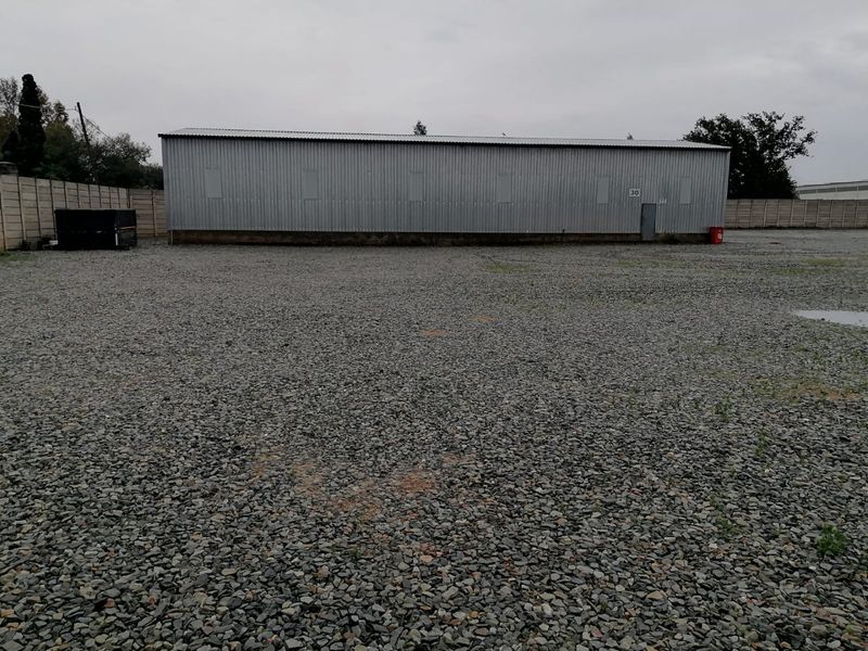 Industrial warehouse building featuring corrugated metal cladding situated on a large, open gravel yard with perimeter walling.