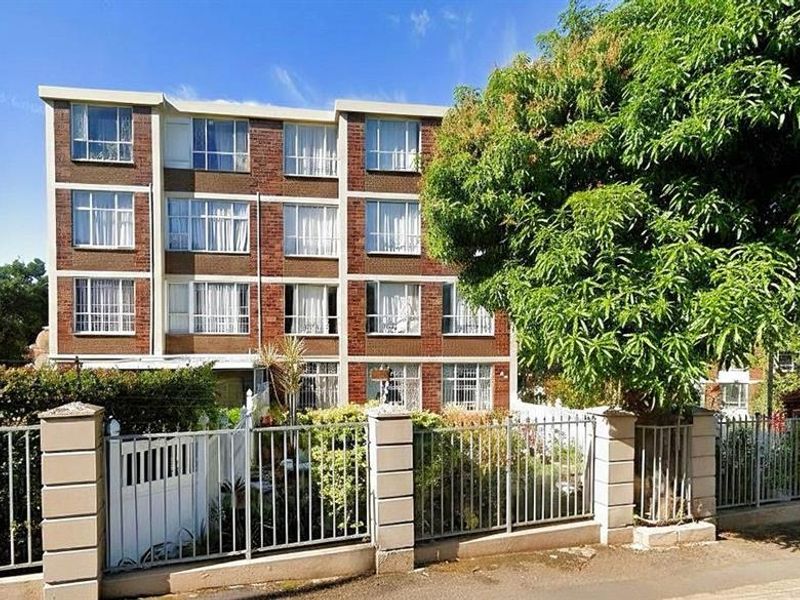 Street-level view of a multi-story brick apartment building featuring metal security fencing and established garden landscaping.
