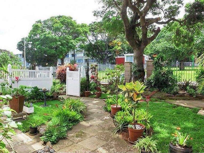 A well-maintained private garden featuring a paved stone walkway, mature trees, and a secure white picket fence with a pedestrian gate.