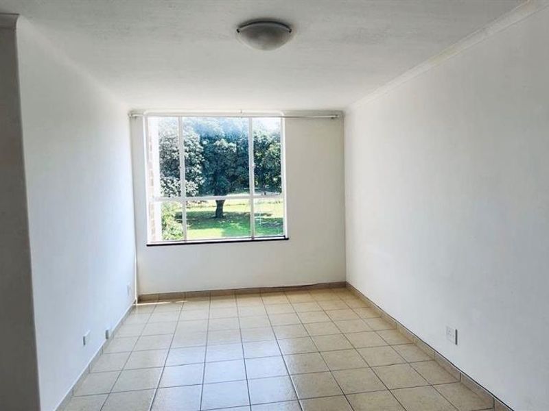 An empty, sunlit living area featuring neutral tiled flooring and a large window with views of the surrounding greenery.