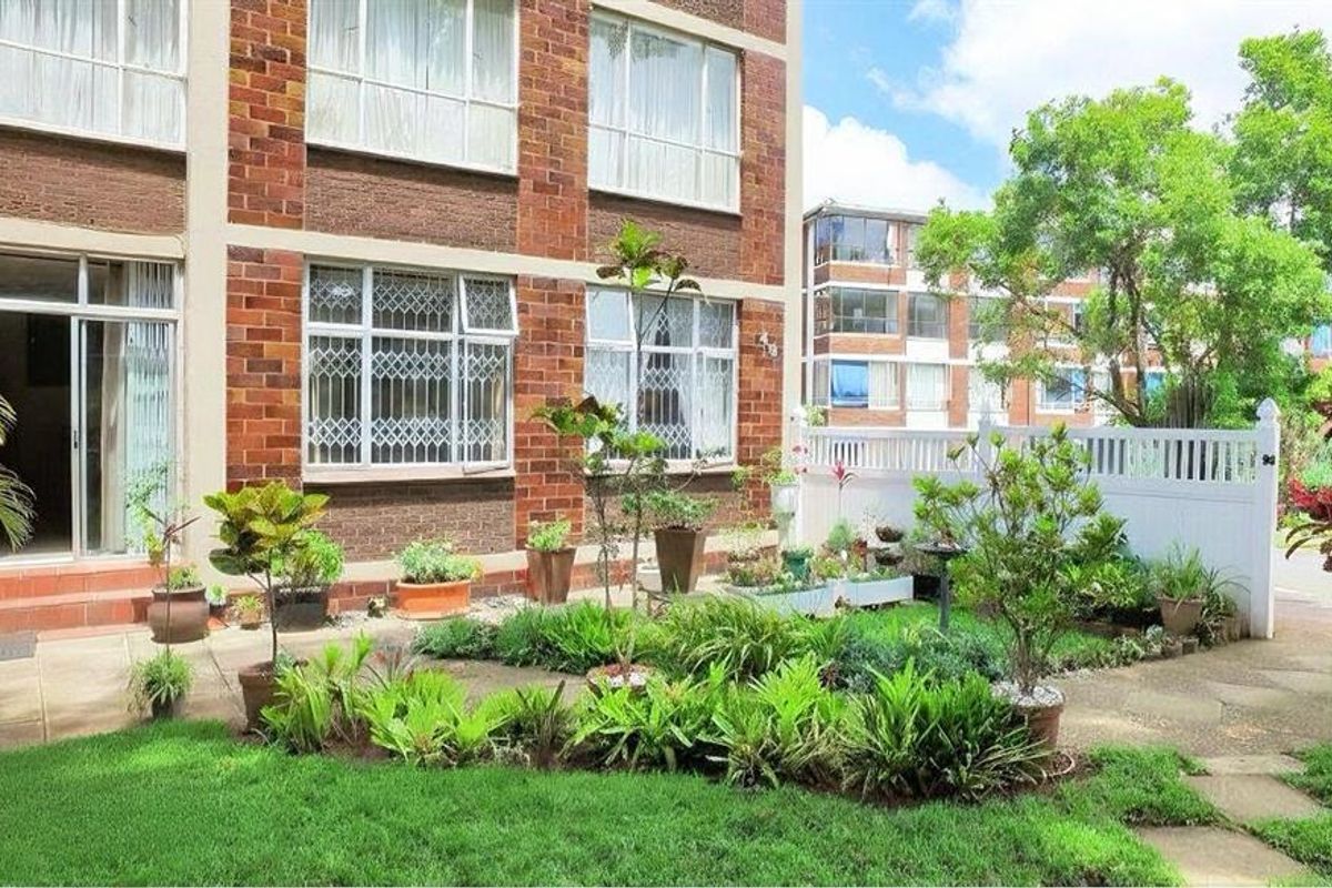 A ground-floor apartment exterior featuring a brick facade, a paved patio area, and a private garden space enclosed by a white picket fence.