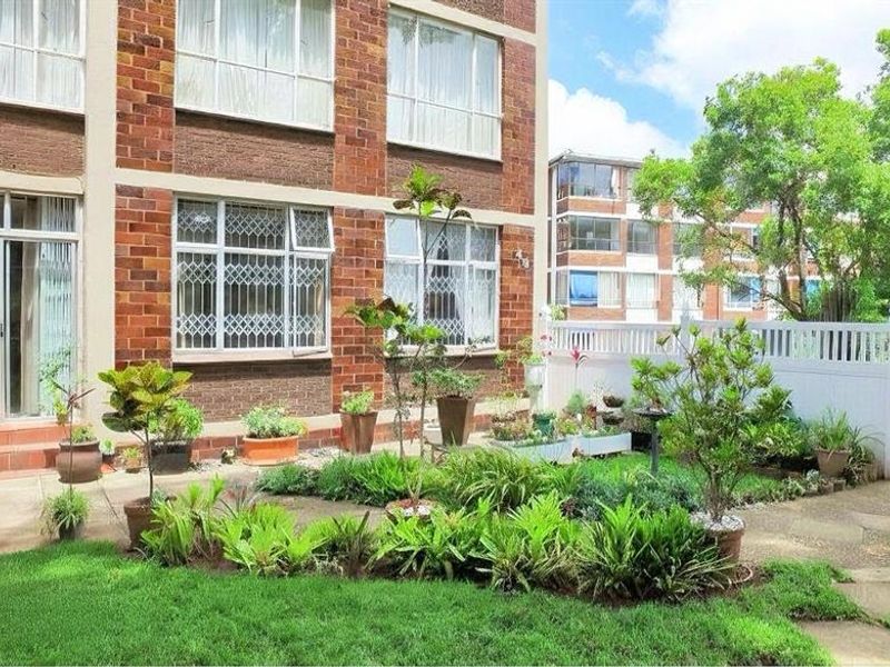 A ground-floor apartment exterior featuring a brick facade, a paved patio area, and a private garden space enclosed by a white picket fence.