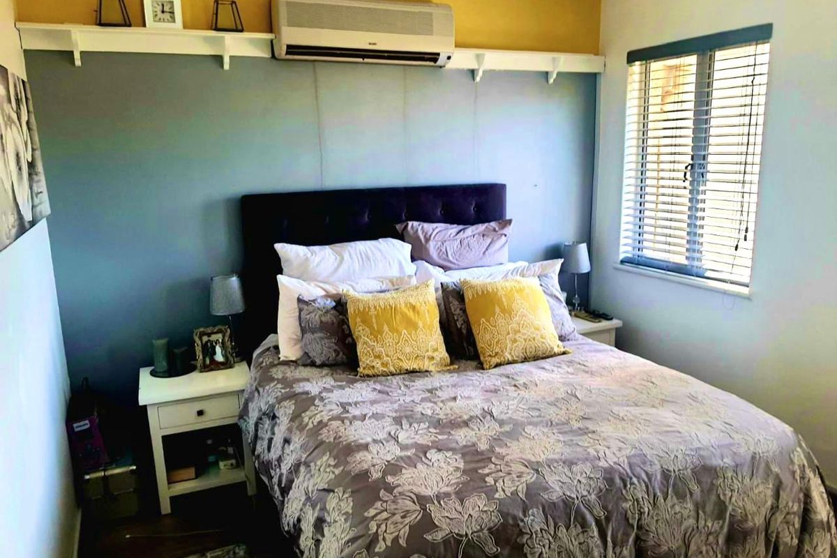 A cozy bedroom featuring a wall-mounted air conditioning unit, a decorative shelf, and venetian blinds for light control.