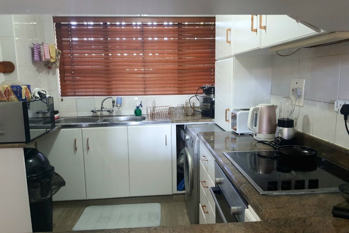 A functional kitchen featuring stone countertops, built-in cabinetry, an electric hob, oven, and integrated washing machine, finished with a tiled backsplash and venetian blinds.