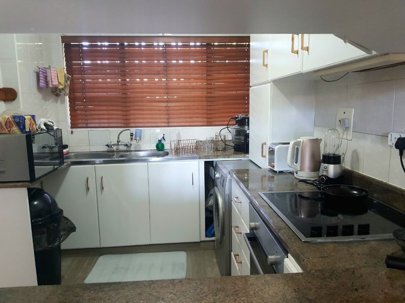 A functional kitchen featuring stone countertops, built-in cabinetry, an electric hob, oven, and integrated washing machine, finished with a tiled backsplash and venetian blinds.