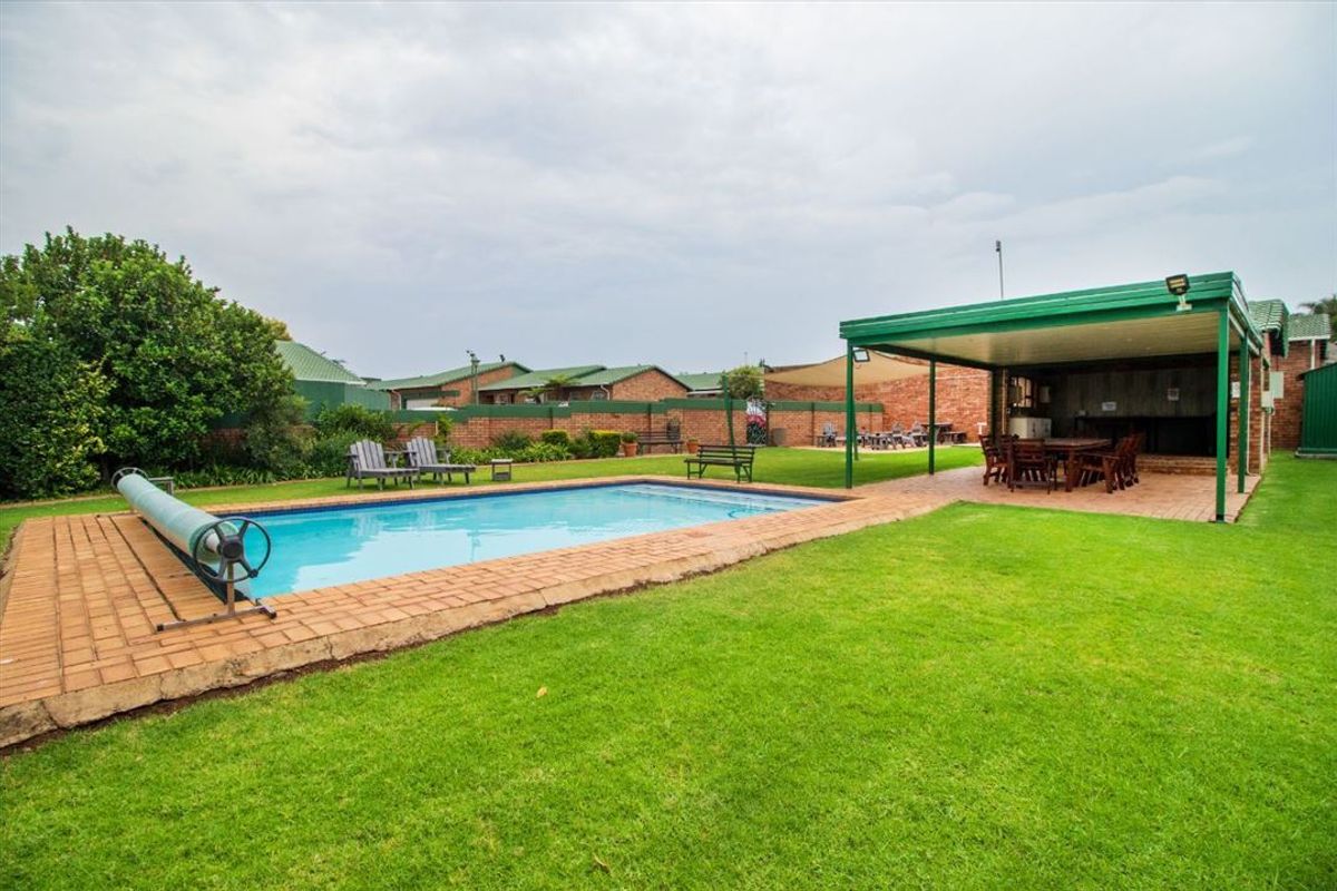 A well-maintained outdoor pool area featuring a swimming pool, surrounding brick paving, a spacious lawn, and a covered patio area.