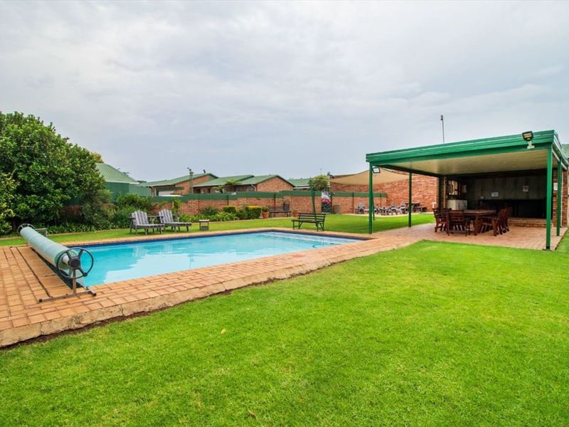 A well-maintained outdoor pool area featuring a swimming pool, surrounding brick paving, a spacious lawn, and a covered patio area.