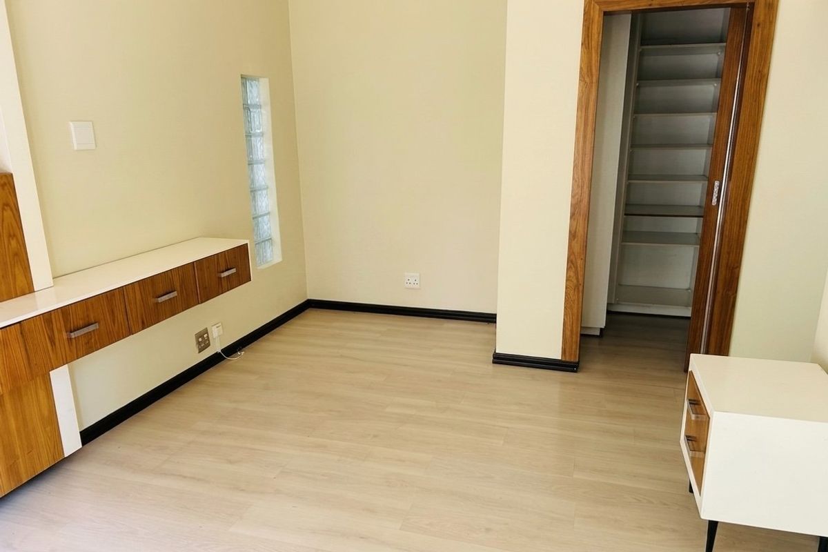 A clean, empty bedroom featuring light laminate flooring, built-in wooden cabinetry, and a unique glass block window detail.