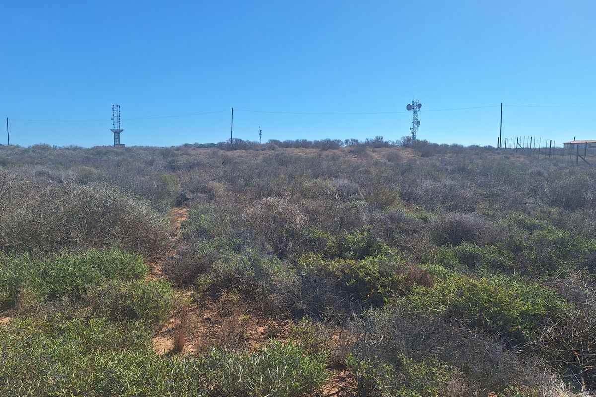 A view of the vacant residential land featuring native scrub vegetation, perimeter fencing, and distant telecommunication towers under a clear blue sky.