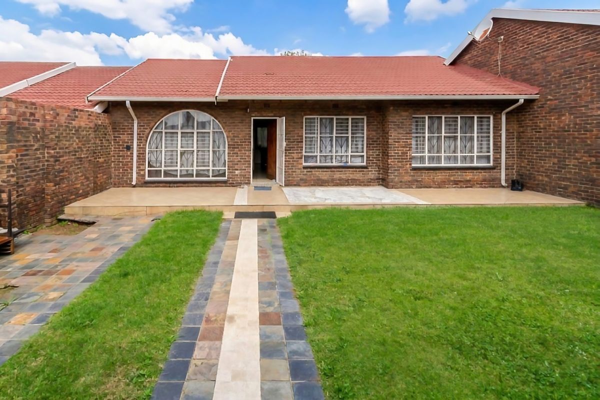 Rear view of a brick townhouse featuring a tiled roof, arched window, paved patio area, and a well-maintained garden lawn.
