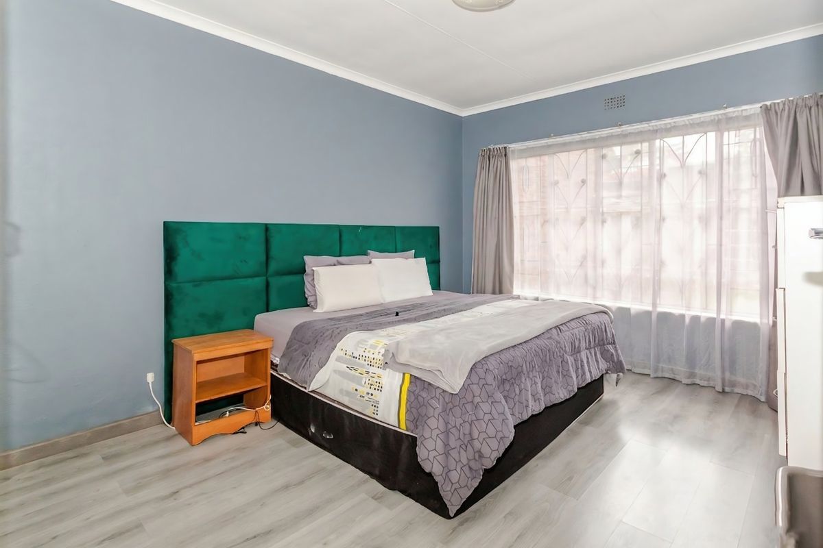 A well-maintained bedroom featuring light-toned laminate flooring, neutral wall paint, and a large window fitted with a security gate.
