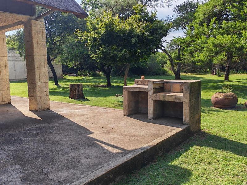 Covered outdoor patio featuring a thatched roof, stone pillars, and a built-in concrete braai area overlooking a spacious garden.
