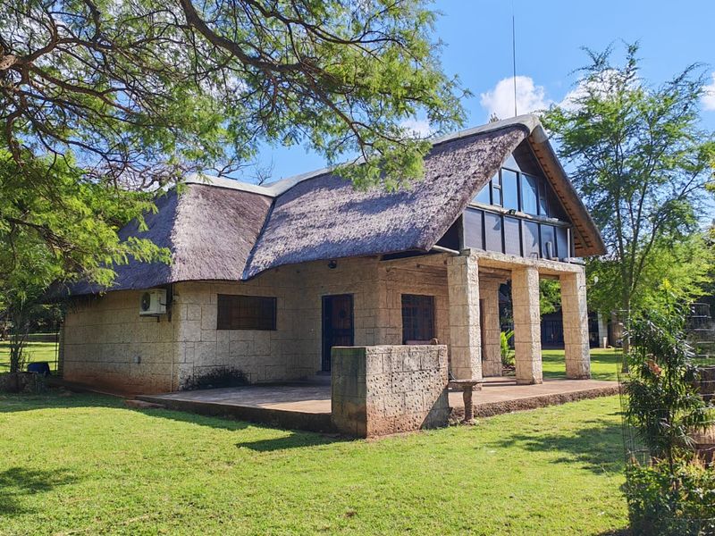 A charming farm-style cottage featuring a traditional thatch roof, stone masonry construction, and a covered patio set within a spacious, well-maintained garden.