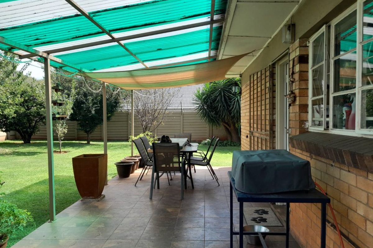 Covered outdoor patio area featuring tiled flooring, brick wall construction, and a green corrugated roof, providing a functional space for outdoor dining and relaxation.