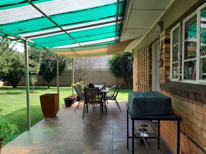 Covered outdoor patio area featuring tiled flooring, brick wall construction, and a green corrugated roof, providing a functional space for outdoor dining and relaxation.