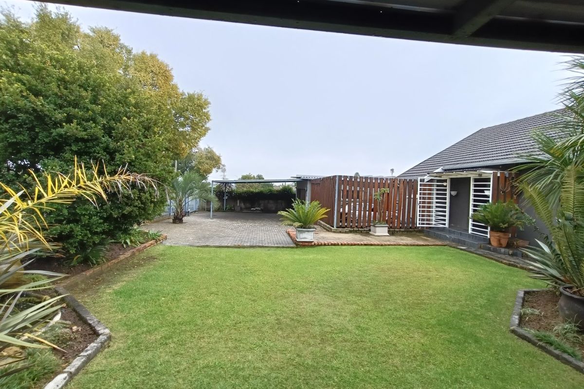 A well-maintained rear garden featuring a manicured lawn, paved driveway area, timber fencing, and a covered carport structure.
