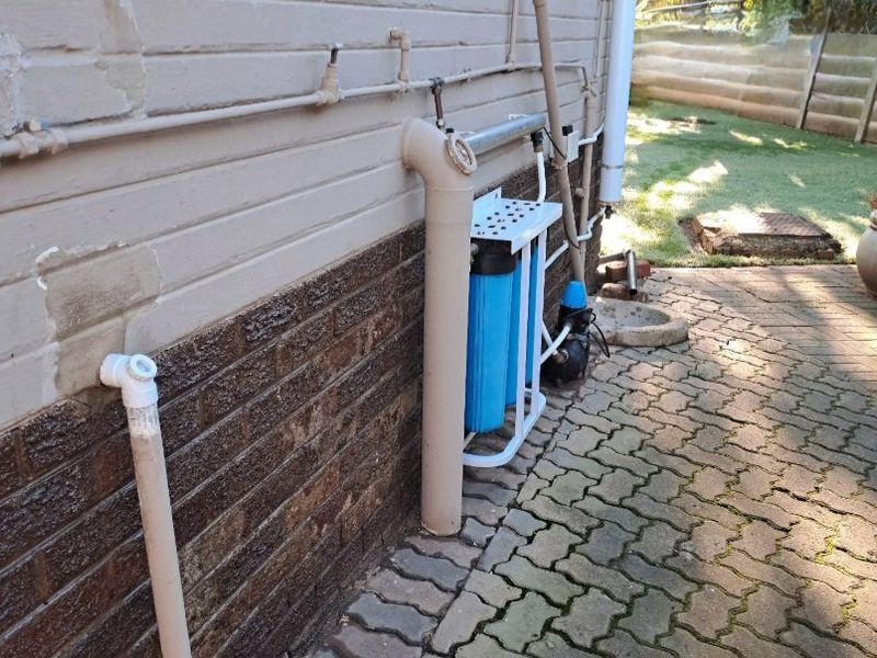 Exterior rear view showing a water filtration system and pump installed against the brick wall with paved ground surface.