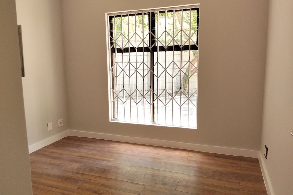 A clean, empty bedroom featuring modern laminate flooring, neutral walls, and a window fitted with a security gate.