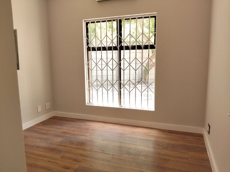 A clean, empty bedroom featuring modern laminate flooring, neutral walls, and a window fitted with a security gate.