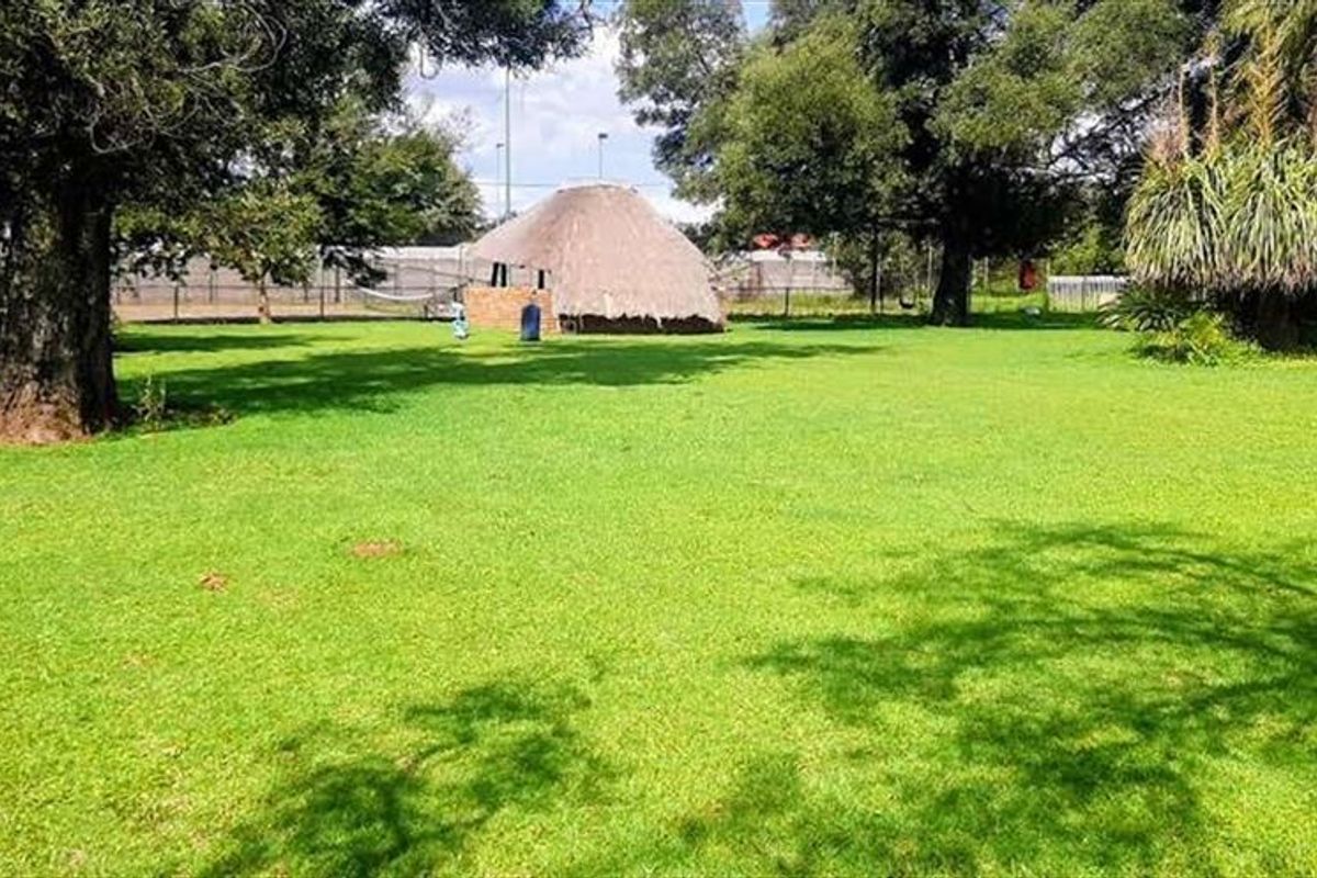 A spacious, well-maintained garden area featuring a traditional thatched lapa structure and mature trees on a large farm property.