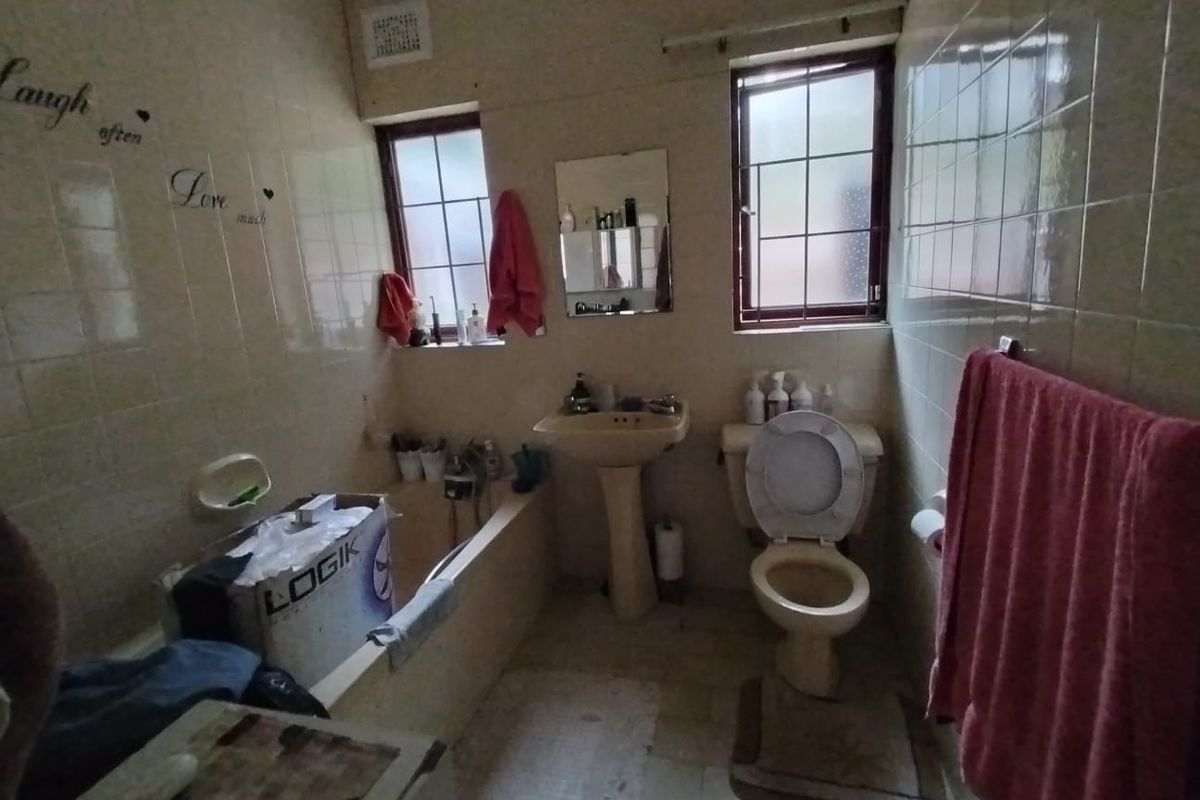 A dated bathroom featuring a pedestal washbasin, toilet, and bathtub with full-height wall tiling and security bars on the windows.