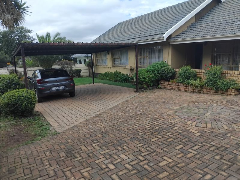 Front exterior view of the house featuring a paved driveway, a covered carport, and established garden beds with brick retaining walls.