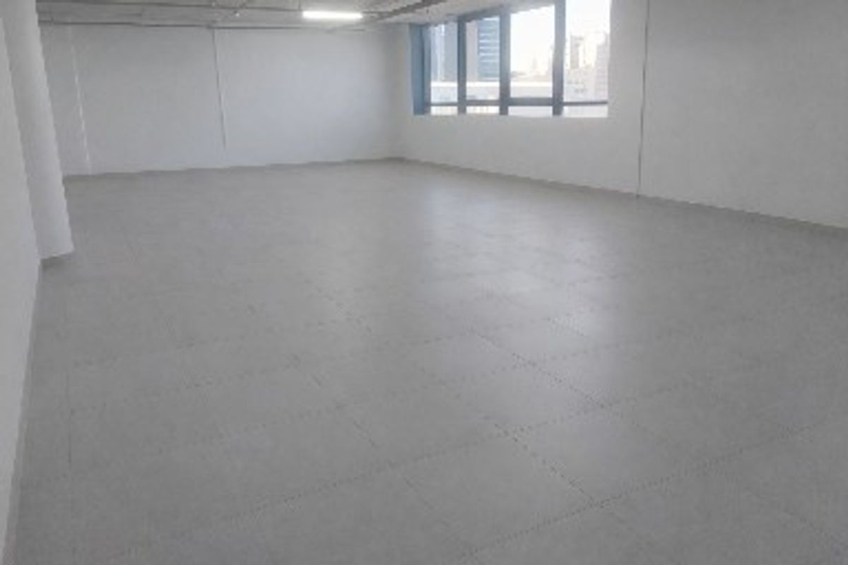 A spacious, empty commercial office area featuring neutral tiled flooring, large windows providing natural light, and a clean, open-plan layout.