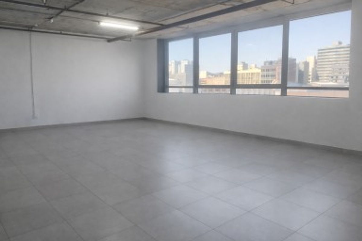 A bright, open-plan commercial office space featuring large windows with city views, tiled flooring, and an exposed concrete ceiling.