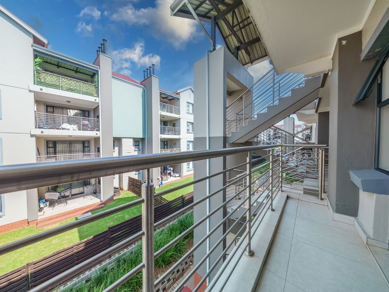 A spacious covered balcony featuring a modern stainless steel balustrade, tiled flooring, and views overlooking the apartment complex grounds and external staircase.
