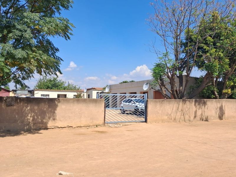 Front view of the property featuring a perimeter wall, metal driveway gate, and satellite dishes, set on an unpaved ground surface.