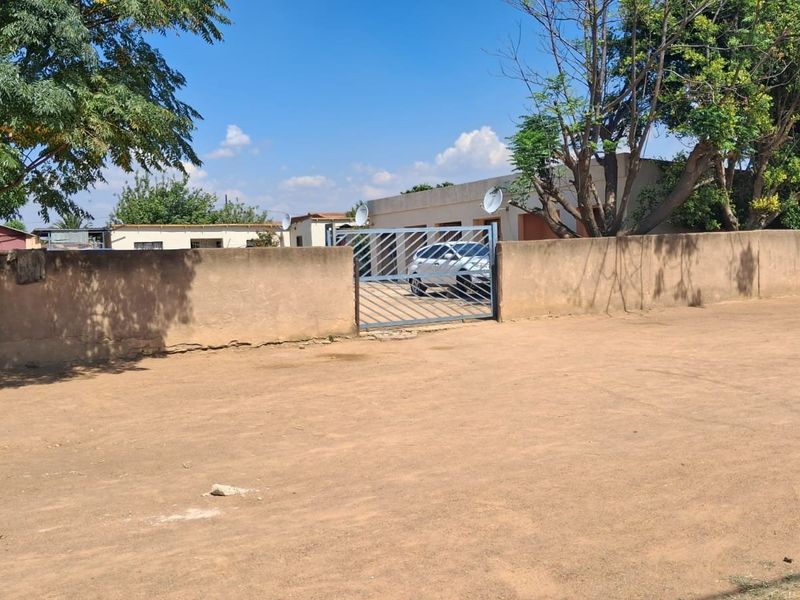 Front view of the property featuring a perimeter wall, metal entrance gate, and a large unpaved driveway area.