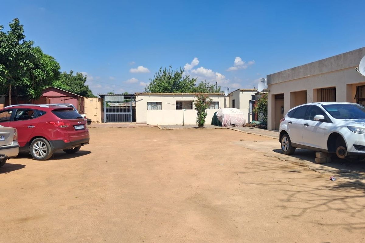 A spacious rear exterior area featuring a large unpaved driveway, concrete pathways, and multiple building access points with satellite dish installations.