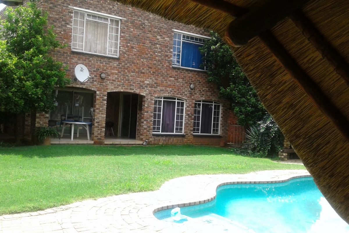 Rear view of a brick house featuring a swimming pool, paved surround, and a thatched lapa structure in the garden.