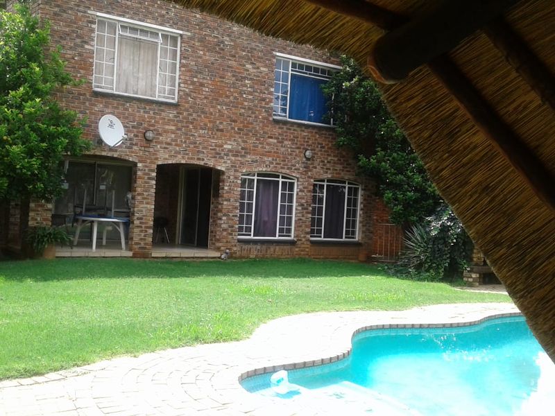 Rear view of a brick house featuring a swimming pool, paved surround, and a thatched lapa structure in the garden.