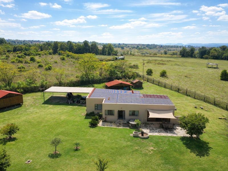 Aerial view of a farm property featuring a main dwelling with a metal roof, attached carport, stone patio, and multiple outbuildings set on expansive grounds.