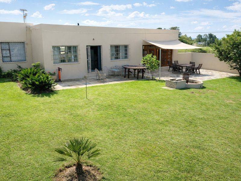 Rear view of the farmhouse featuring a stone-paved patio, shade sail, and a built-in brick fire pit overlooking a spacious lawn.